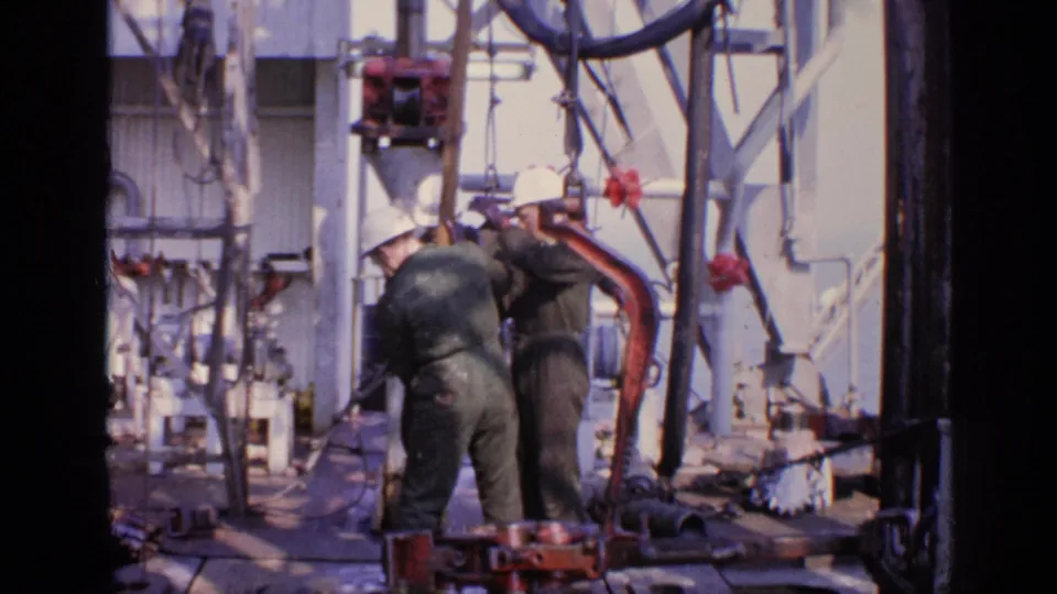 1970 United Kingdom 8mm frame showing two oil rig workers bending over red equipment on a cluttered offshore platform deck.