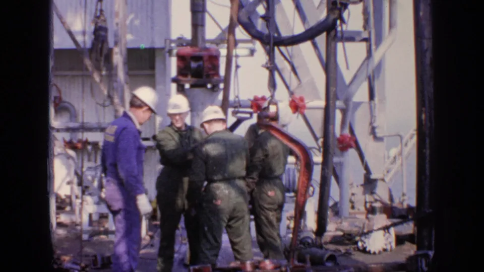 Hard-hatted workers converse near exposed pipes and rig equipment in 1970 United Kingdom home-movie footage.