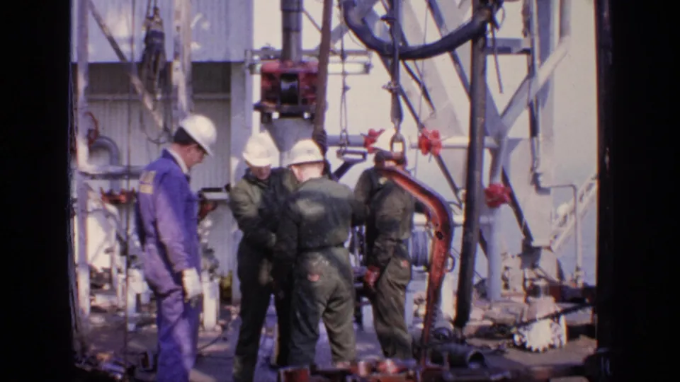 Workers in hard hats and coveralls stand near drilling-site machinery, pipes, and cables in 1970 United Kingdom footage.