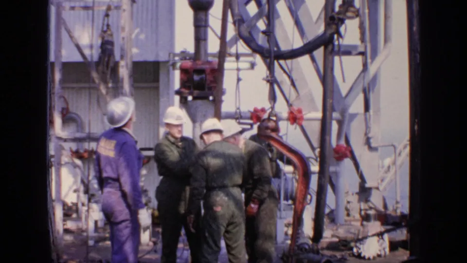 A small crew in hard hats stands among pipes, cables, and oil rig or drilling-site equipment in 1970 United Kingdom footage.
