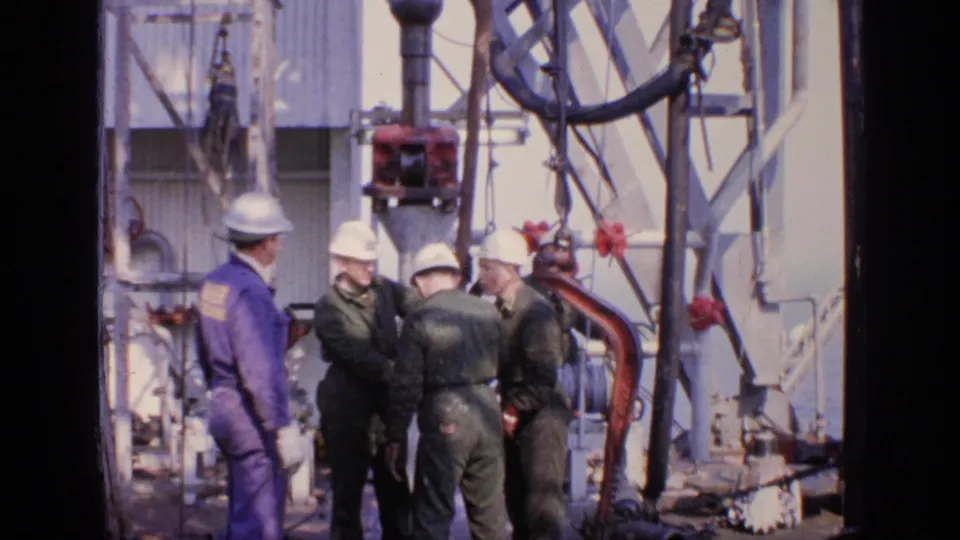 Four hard-hatted workers in green and blue coveralls gather around offshore rig equipment in 1970s United Kingdom footage.