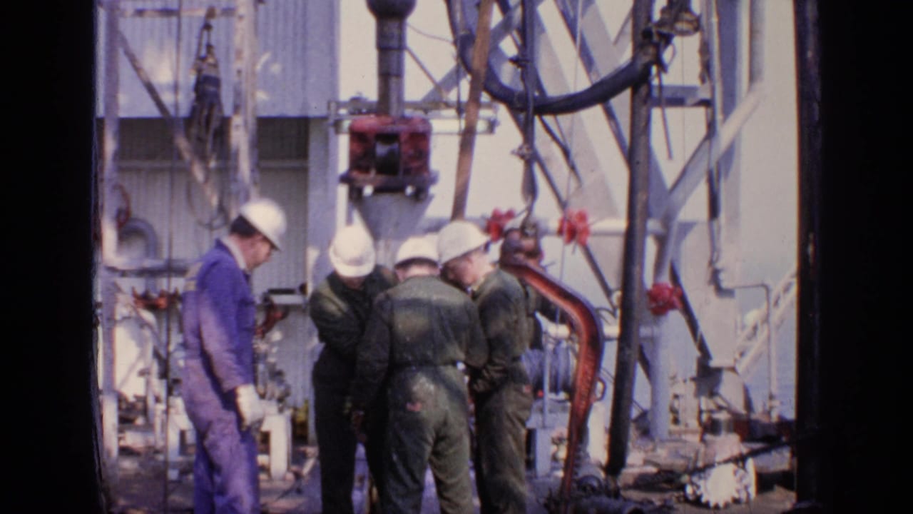 Workers in hard hats look down at equipment on an oil rig deck.