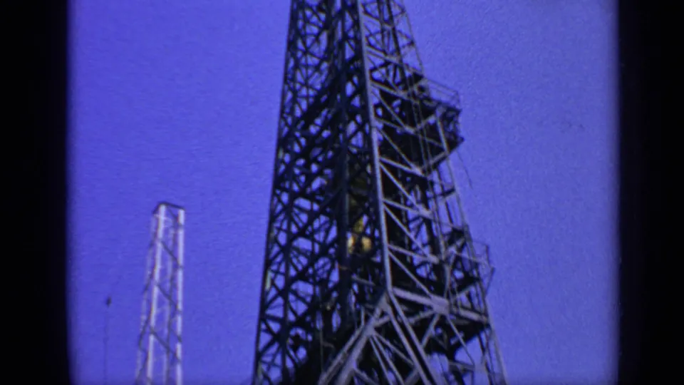 1970 United Kingdom frame showing the steel tower of an offshore oil rig against blue sky, with 8mm film-edge shadows.