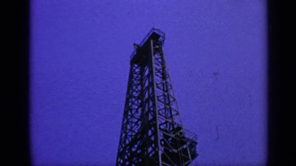 1970 United Kingdom offshore rig derrick framed by 8mm film borders, with lattice steelwork silhouetted against blue sky.