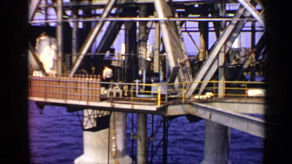 1970 United Kingdom 8mm view of an offshore platform deck with machinery, railings, sea below, and a worker visible in a white shirt.