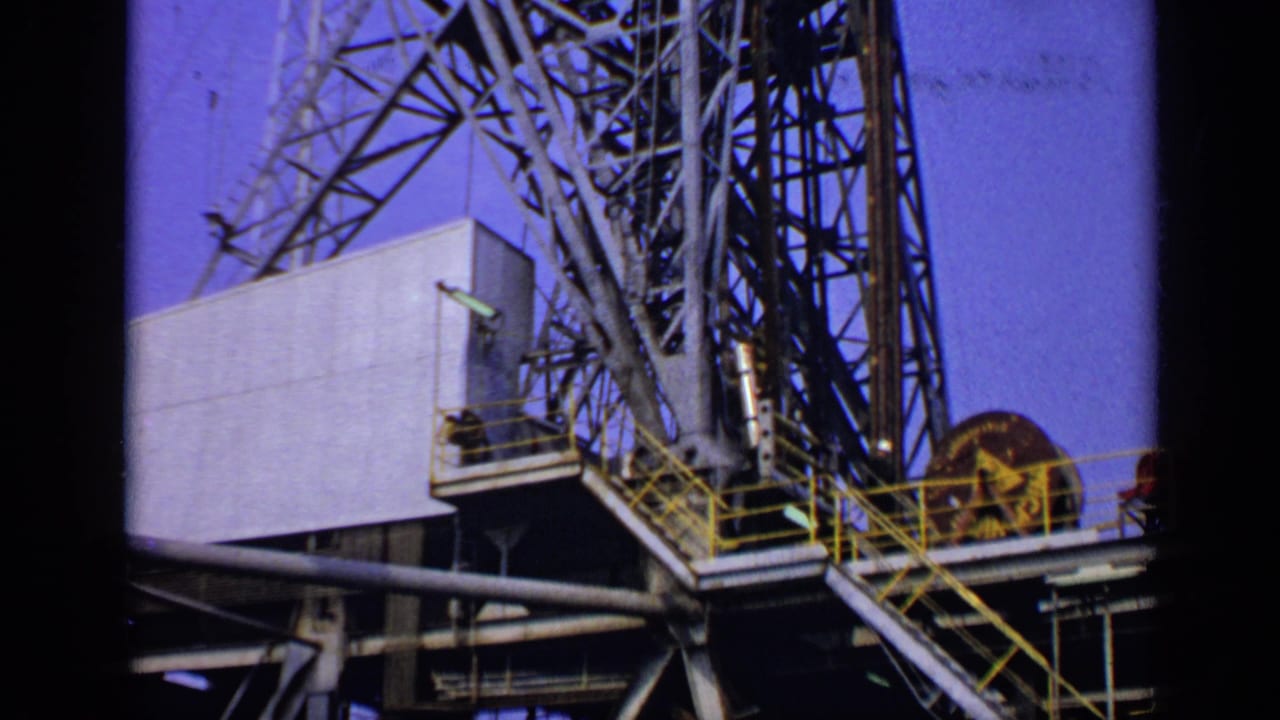 1970 United Kingdom industrial footage frame with steel derrick sections, a platform, yellow railings, and a large spool against the sky.