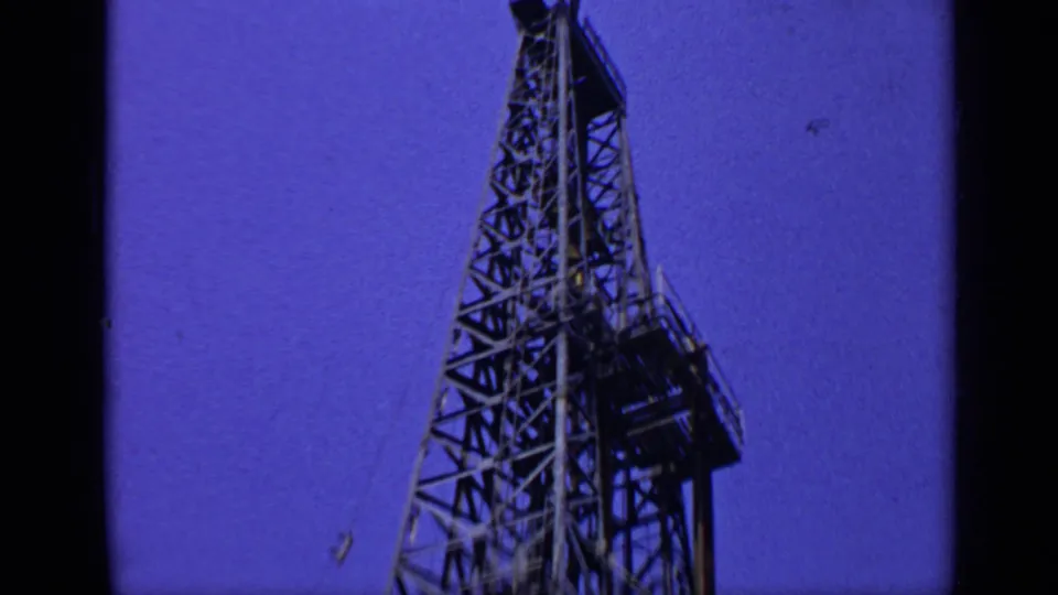 1970 United Kingdom 8mm frame looking up at a tall steel derrick tower with platforms and latticework visible.