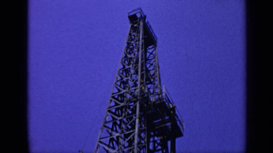 1970 United Kingdom 8mm frame showing a refinery derrick tower from below, with lattice steelwork and platform elements.