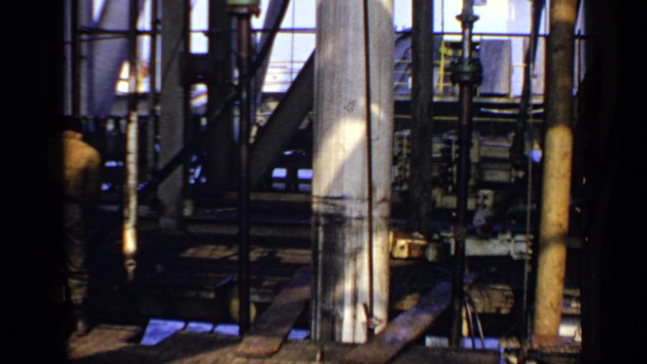 Oil rig interior with pipes, beams, machinery, and a partly visible worker in the background.