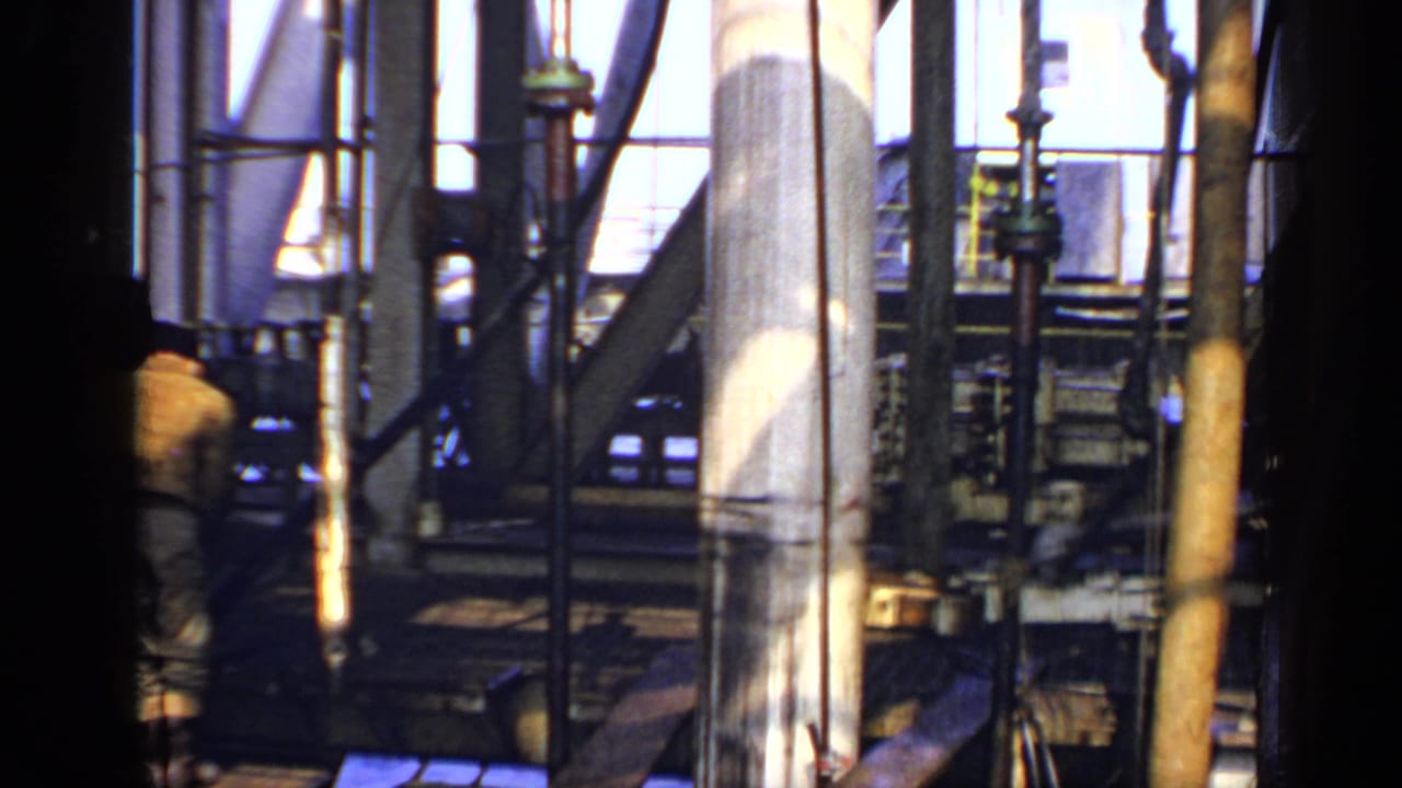 Rig machinery and vertical pipes with a worker partly obscured behind dark foreground structure.