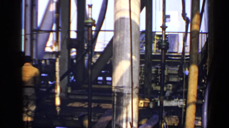 1970 United Kingdom rig interior filmed on 8mm, showing structural steel, machinery, pipes, and a blurred worker moving near the edge.