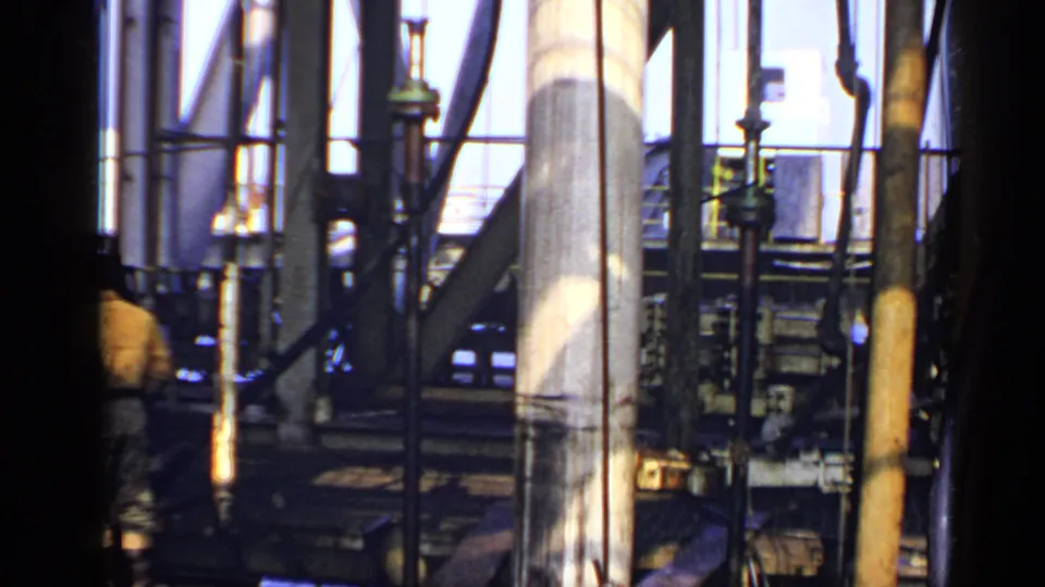 United Kingdom oil-rig interior from 1970, with 8mm grain, bright window light, steel structure, machinery, and a partly visible worker.