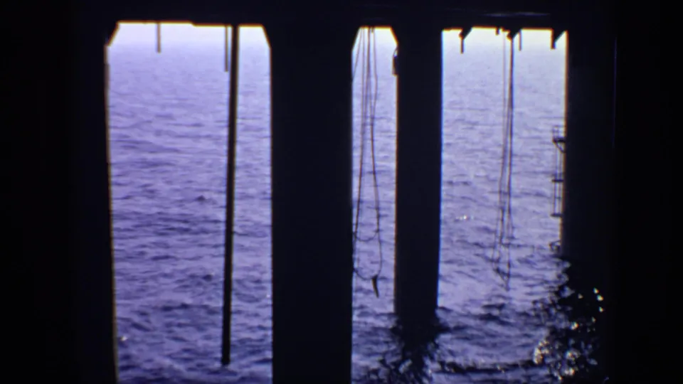 1970 8mm coastal frame with pier pillars, hanging ropes or fishing lines, and blue-purple sea visible through the underside structure.