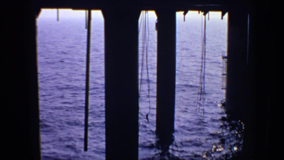 Dark pier columns and suspended ropes frame the calm sea in this 1970 United Kingdom home-movie still.