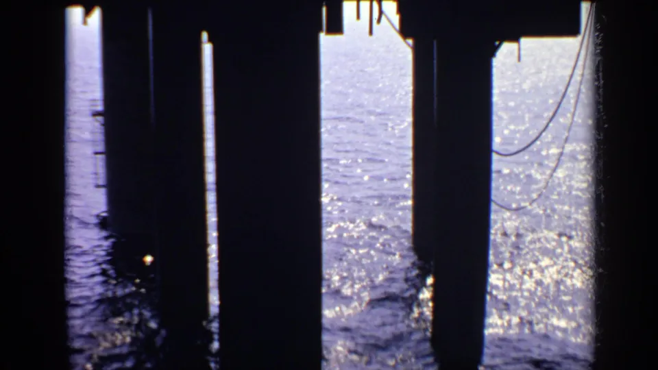 1970 UK 8mm frame showing sunlit sea through dark pier supports, with high contrast between shadowed columns and bright water.
