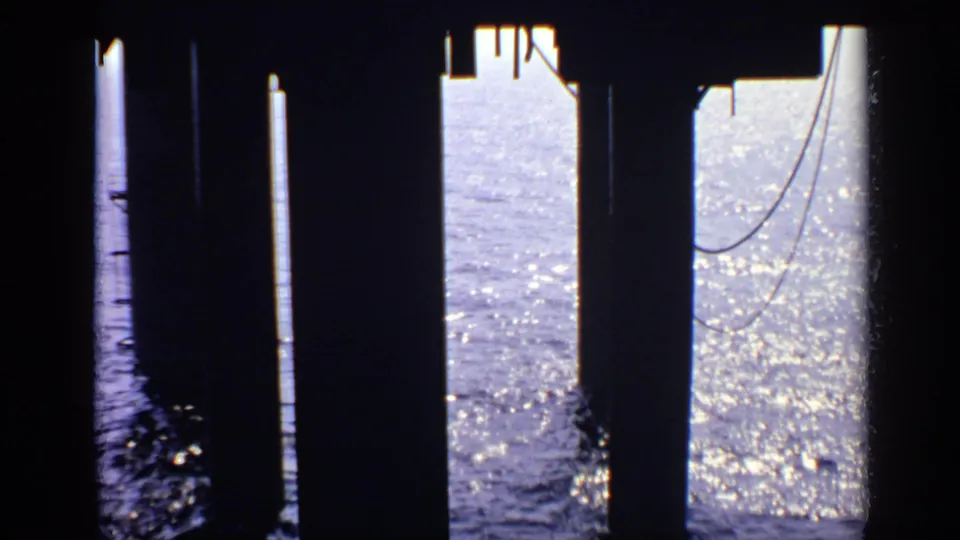 Shadowed pier structure with concrete pillars and hanging cable-like lines silhouetted against bright, reflective sea water.