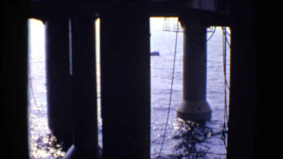 1970 UK coastal frame with shadowed pier columns, a visible concrete base, and bright blue sea beyond the structure.