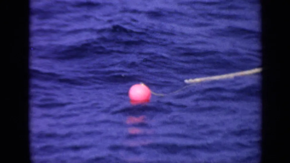 1970 United Kingdom 8mm frame of a red-pink buoy floating low on blue water, with its pale tether line visible nearby.