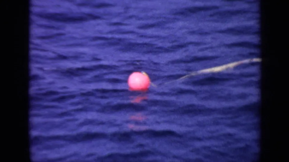 Close 1970 United Kingdom 8mm view of the red-pink buoy and tether line on dark blue rippling water.