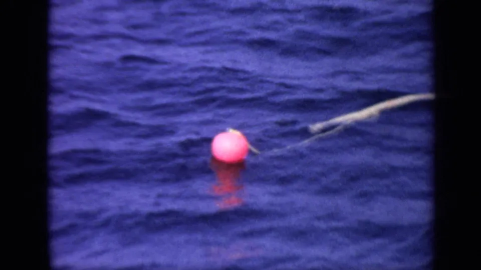 1970 United Kingdom 8mm frame with a pink buoy on deep blue water and a pale tether line crossing behind it.