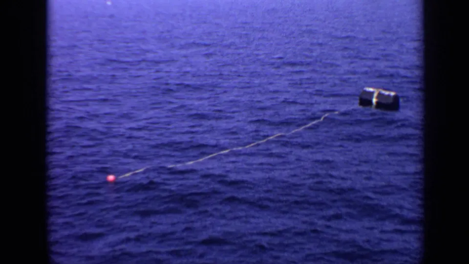 1970 United Kingdom 8mm frame showing a thin line across open water, connecting a small pink float to a dark cylindrical object.