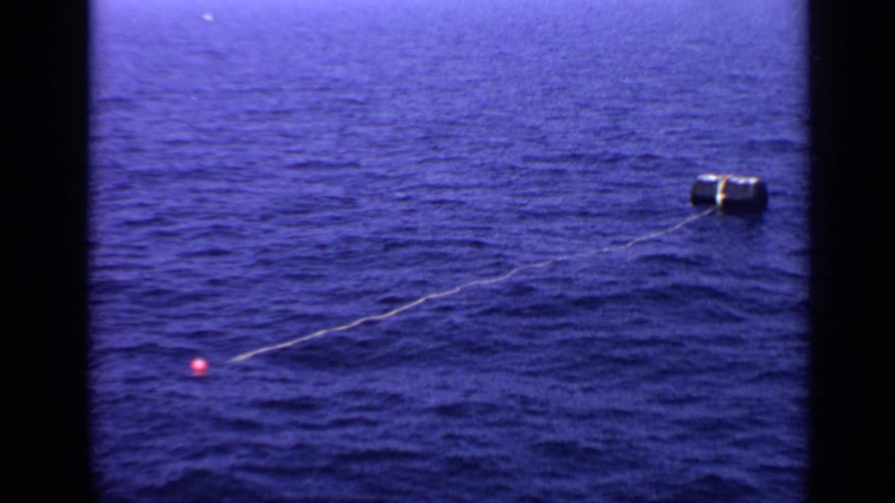 Cylindrical buoy floating in blue ocean with a line leading toward a small red marker.