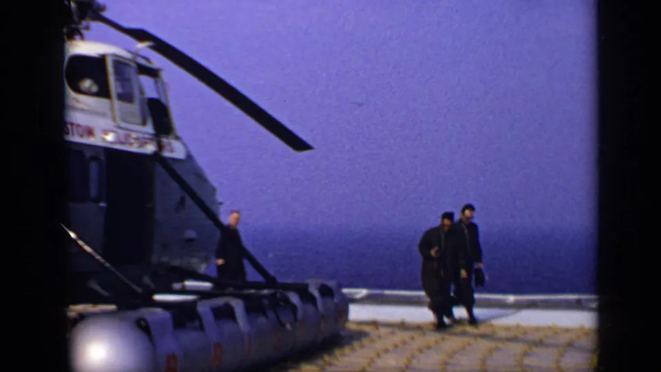 1970 United Kingdom 8mm frame with men near a helicopter marked Bristol Helicopters beside the sea.
