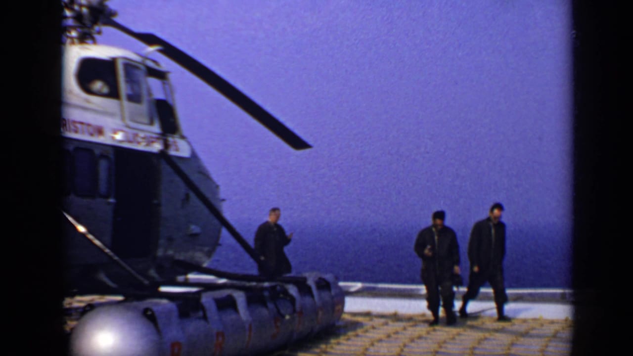 Helicopter on an offshore deck with three men walking nearby and sea behind them.