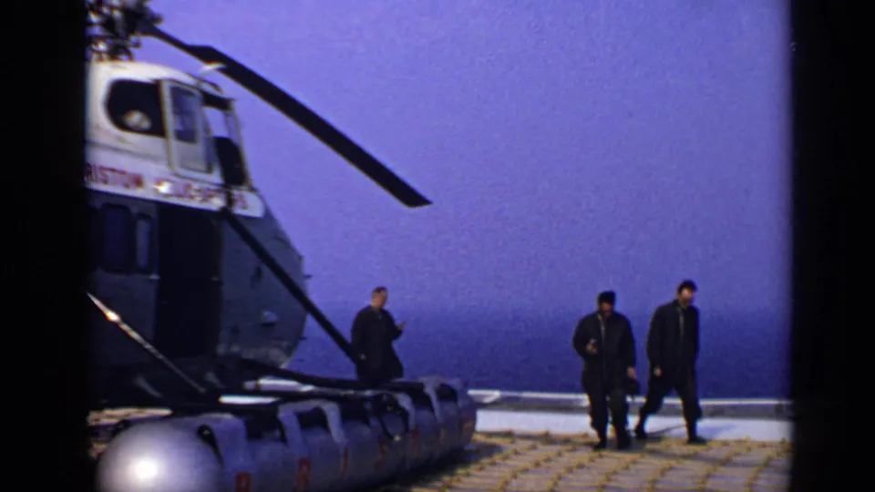 1970 United Kingdom 8mm frame showing a Bristol-marked helicopter on a deck as three adult men walk across the helipad with ocean in the background.