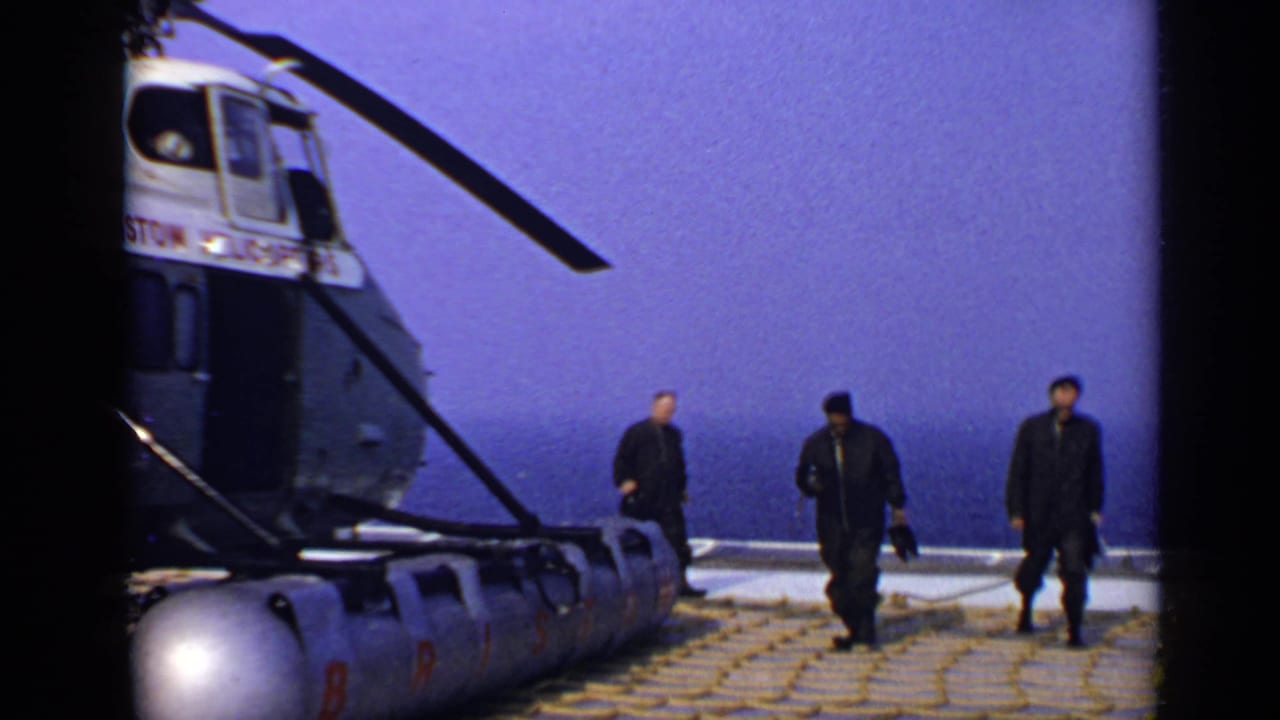 Three men walk across a deck beside a helicopter with the ocean in the background.