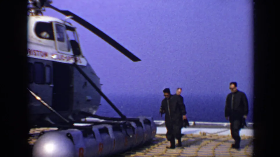 Adult men walk past the nose of a helicopter on an offshore deck, with the hazy ocean horizon and saturated 8mm color visible.