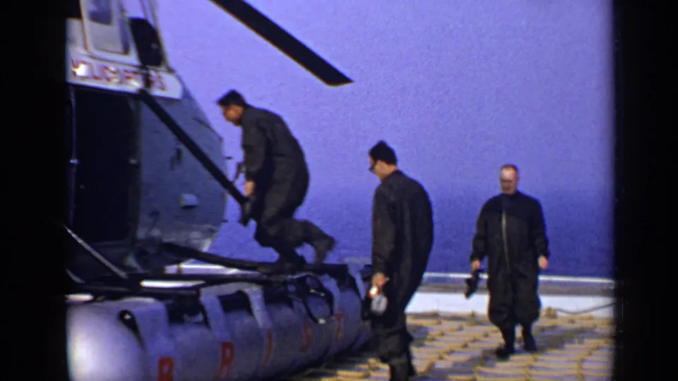 One adult man steps up to board the Bristol-marked helicopter while two others continue across the offshore deck, with sea and sky behind them.