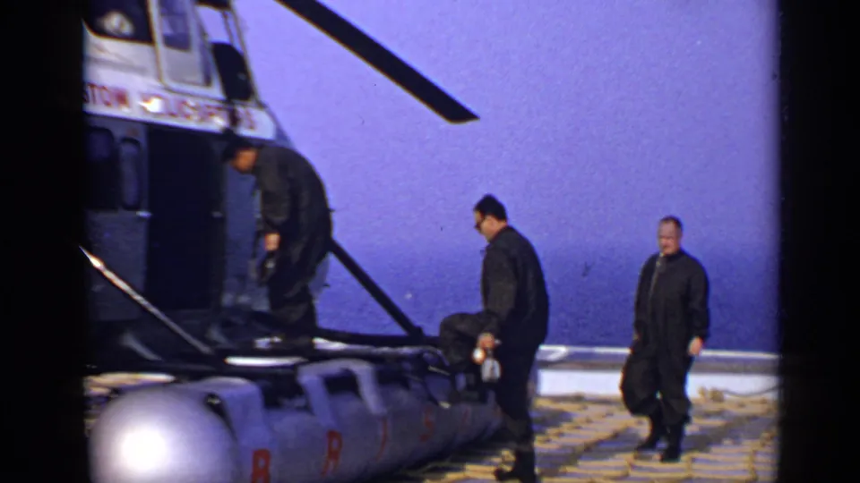 1970 United Kingdom 8mm frame showing three men approaching and boarding a Bristol Helicopters aircraft under a clear sky.