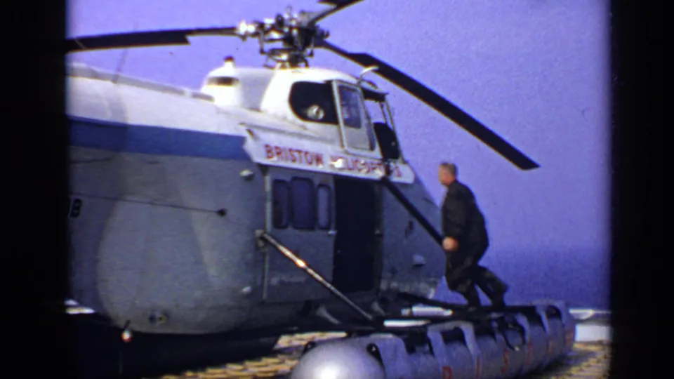 1970s United Kingdom 8mm frame showing a man climbing steps into a Bristow helicopter as the main rotor spins.