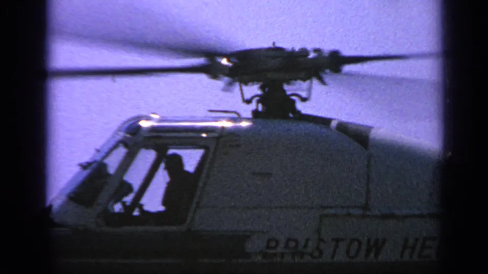 1970s 8mm side view of the Bristow helicopter cockpit, with rotor blur above and a seated figure visible inside.