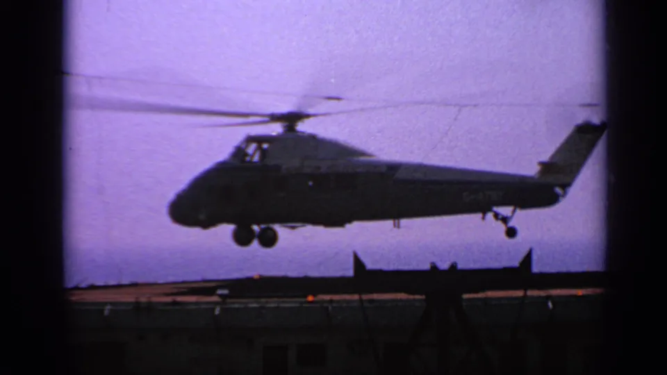 1970 United Kingdom 8mm frame showing the helicopter airborne over the helipad, tail and landing gear visible in silhouette.