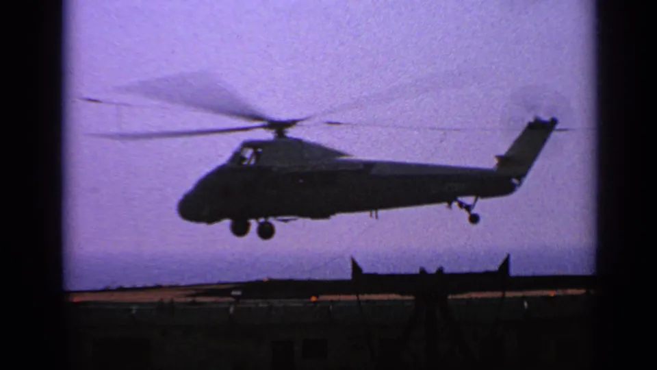 1970 United Kingdom 8mm frame showing a military helicopter lifting from an aircraft carrier deck in silhouette.
