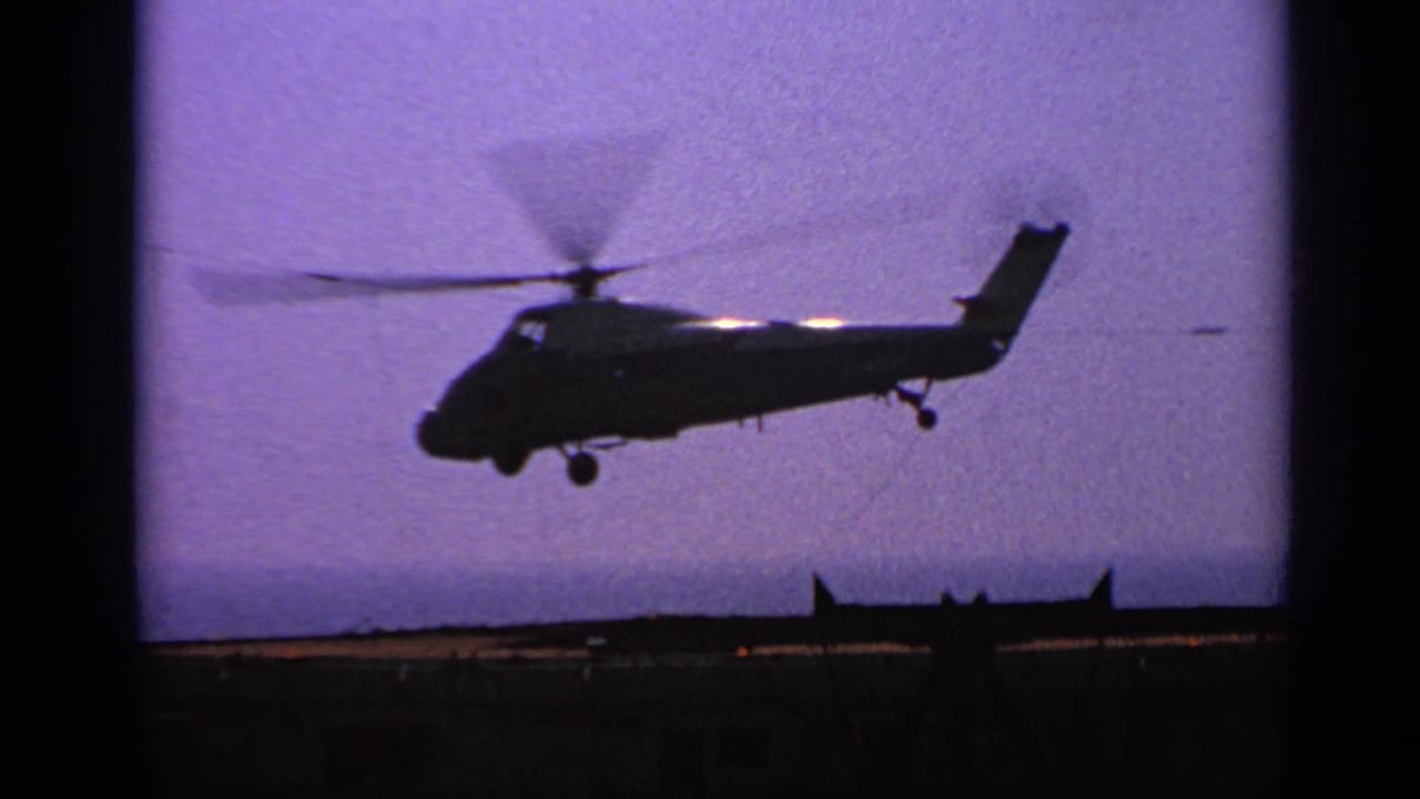A silhouetted helicopter continues its takeoff above a carrier deck, with ship structures low on the horizon.