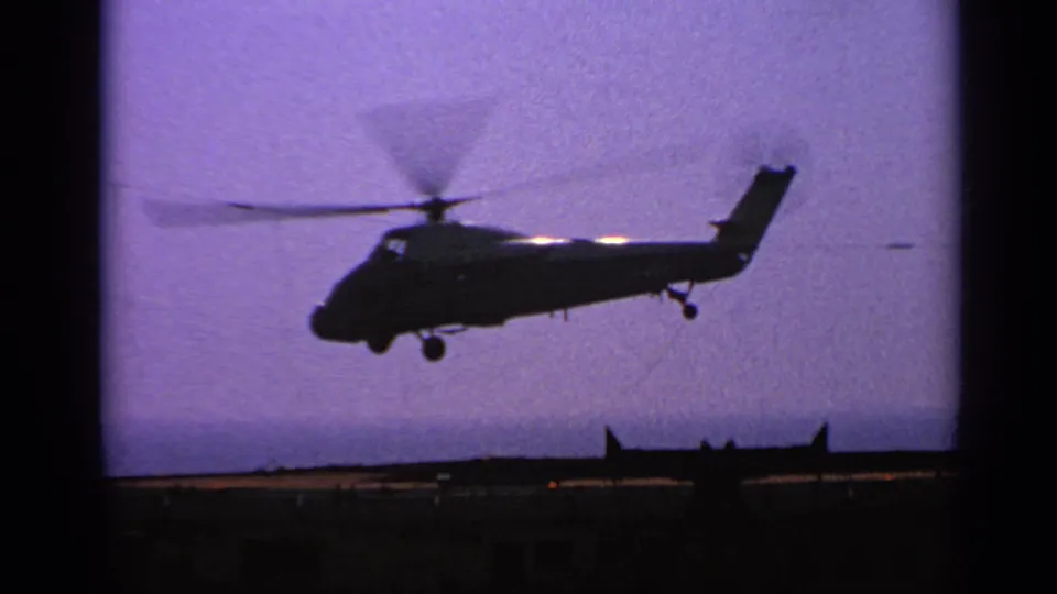 A silhouetted helicopter continues its takeoff above a carrier deck, with ship structures low on the horizon.