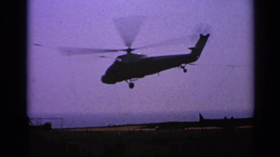1970 8mm frame of a military helicopter climbing away from an aircraft carrier deck under a hazy purple sky.