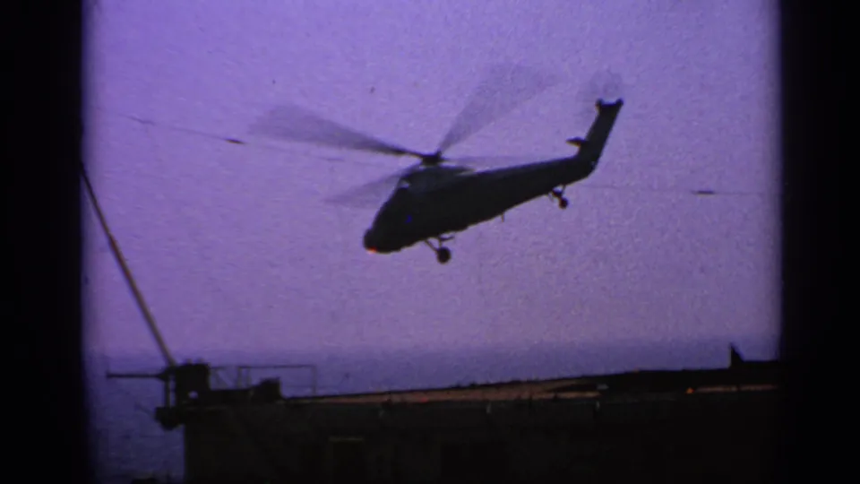 The helicopter climbs away in silhouette, framed by carrier deck details and dark 8mm film borders.