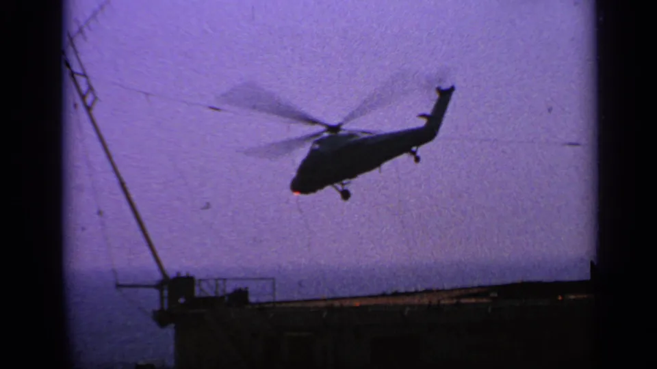 1970 United Kingdom 8mm frame showing a helicopter above a ship deck at sea, with rotor blur against a purple sky.