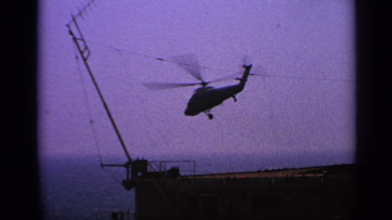 1970 United Kingdom 8mm frame of a helicopter moving away from a ship deck, seen against a purple-blue sky and sea horizon.