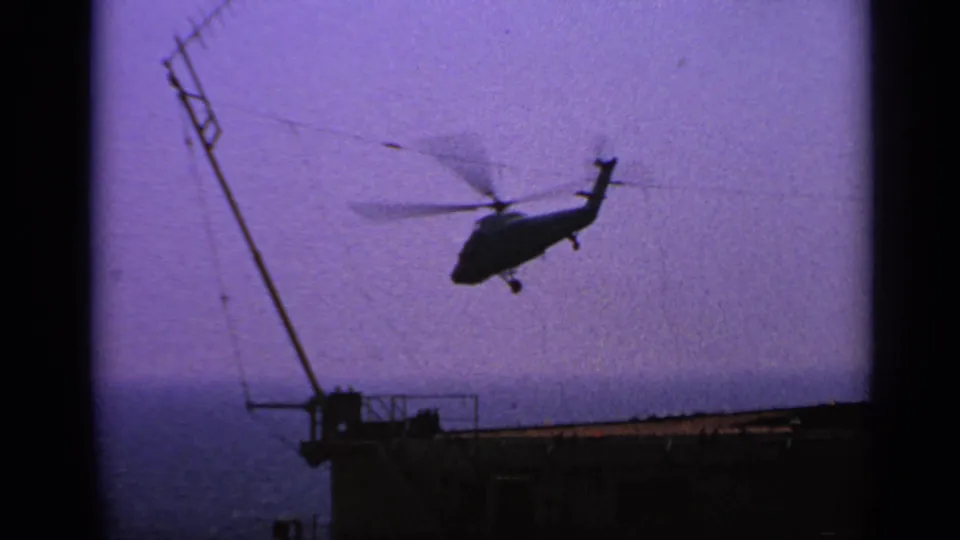 1970 United Kingdom 8mm frame of a helicopter moving away from a ship deck, seen against a purple-blue sky and sea horizon.