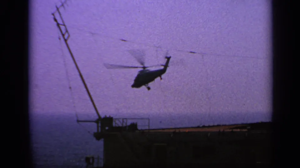 1970 United Kingdom 8mm frame showing a small helicopter in flight near a ship deck, with mast, railing, and ocean visible.