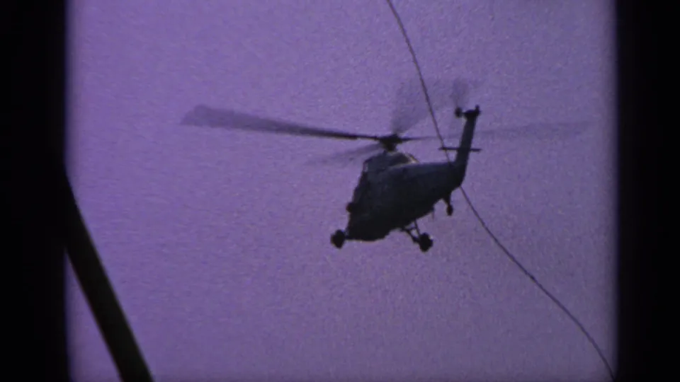 1970 United Kingdom 8mm frame with a closer helicopter silhouette in flight, rotor blur, purple sky, and a dark shipboard line crossing the view.