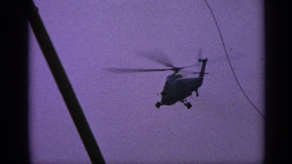 1970 United Kingdom 8mm frame with a distant helicopter silhouette, motion-blurred rotors, and dark foreground framing against a purple sky.