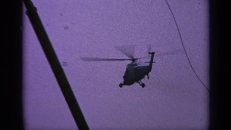 1970 United Kingdom 8mm frame showing the helicopter silhouetted below a dark diagonal support, with purple sky and visible film grain.