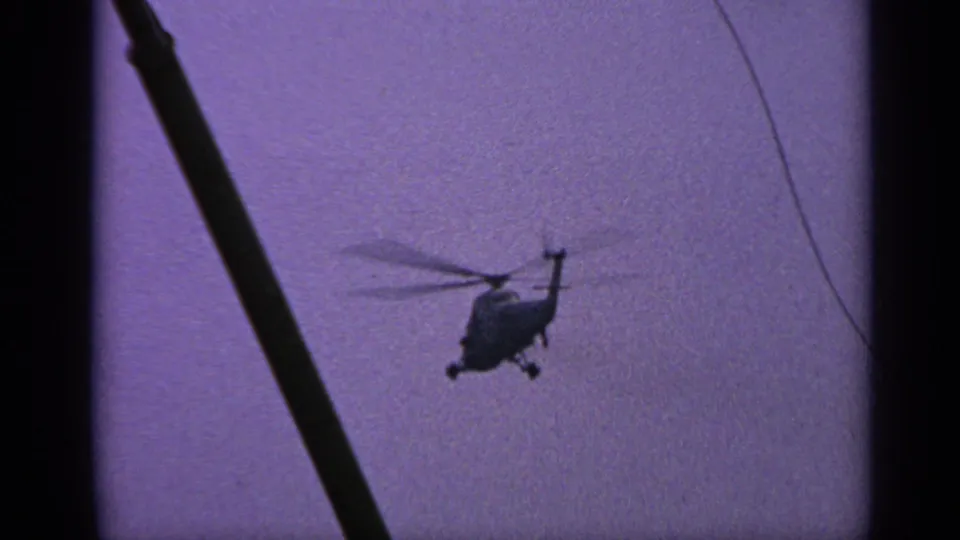 1970s United Kingdom 8mm frame showing a distant silhouetted helicopter hovering or moving across a purple-tinted sky behind a dark pole.