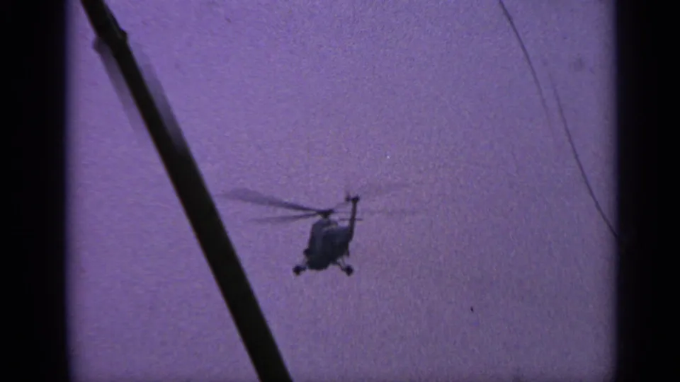 1970s United Kingdom 8mm frame with a small dark helicopter silhouette and spinning rotor visible against grainy purple sky.